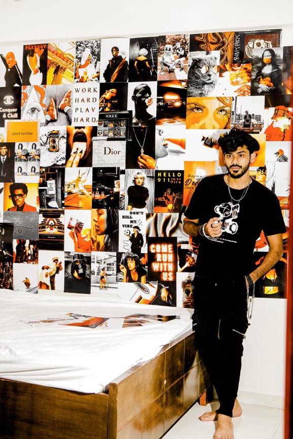 Man in black outfit standing against a wall with orange and black posters collage